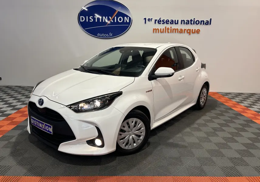 Toyota Yaris hybride blanche vue 3/4 avant droit en intérieur avec phares et antibrouillard allumés.