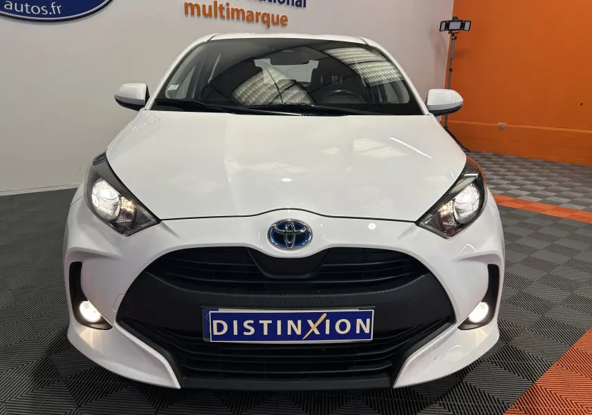 Vue frontale d'une Toyota Yaris blanche 2021 avec phares allumés et plaque Distinxion dans un showroom.
