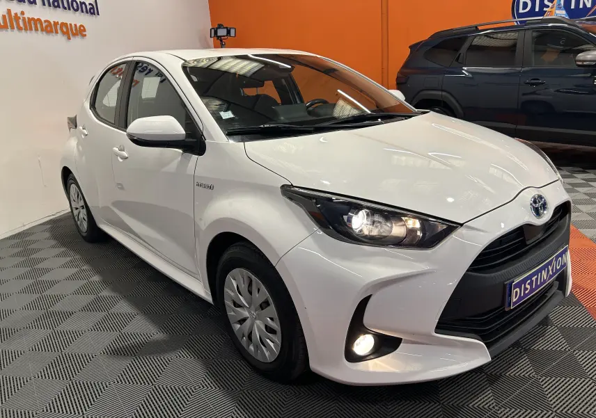 Vue 3/4 avant droite d'une Toyota Yaris 116h blanche avec phares allumés, en intérieur sur sol damier gris.