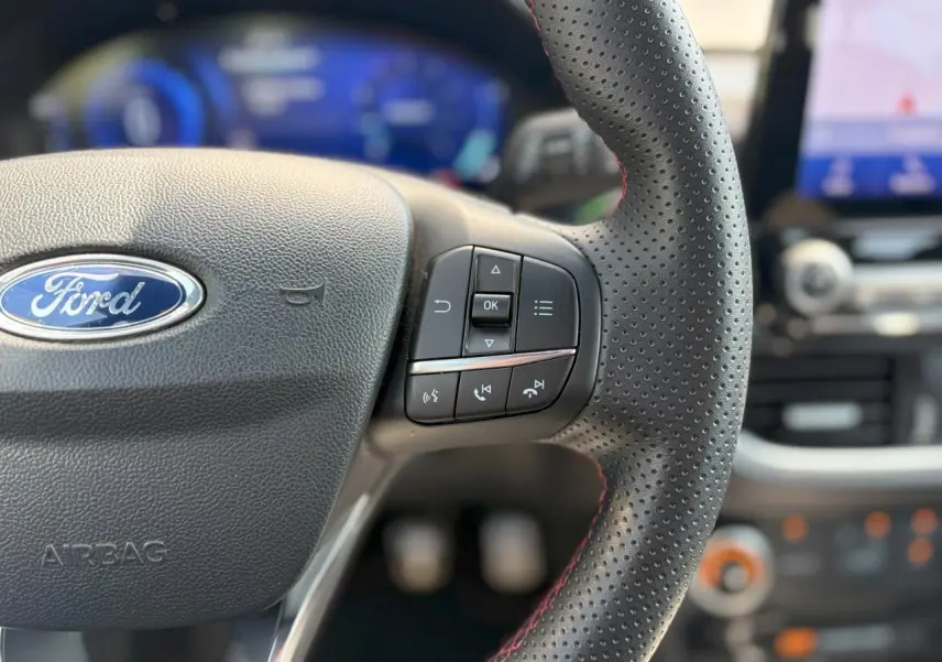 Gros plan sur le volant perforé avec surpiqûres rouges du Ford Puma ST Line, tableau de bord numérique et écran GPS en arrière-plan flou.