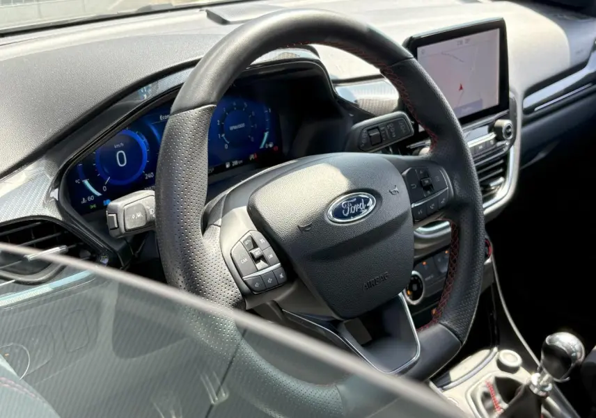 Vue rapprochée du volant noir perforé avec surpiqûres rouges et tableau de bord numérique du Ford Puma Gris Solar 2020.