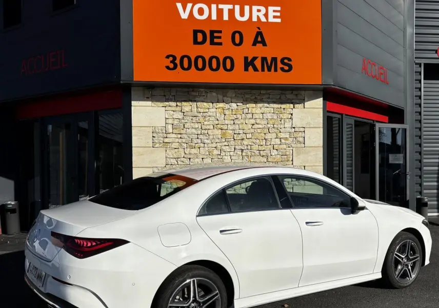 Vue 3/4 arrière droite d'une Mercedes CLA 250 e AMG Line blanche stationnée devant un bâtiment avec panneau orange.