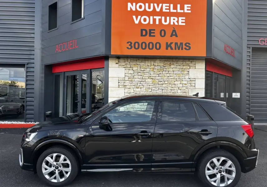 Audi Q2 noir vue de profil côté gauche, garée devant un bâtiment avec panneau orange et gris.