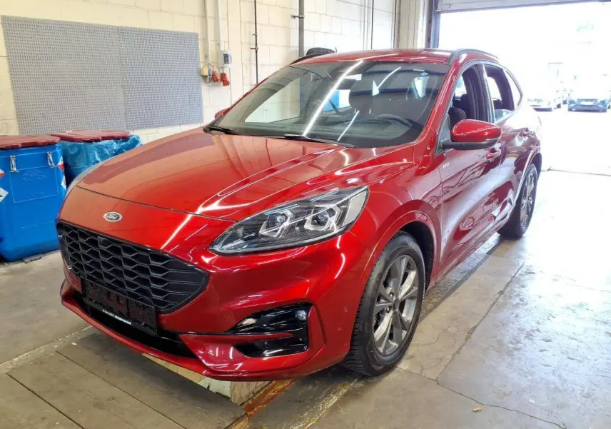 Ford Kuga 2024 rouge Lucifer en 3/4 avant droit, avec calandre noire et jantes alliage visibles en intérieur d'atelier.