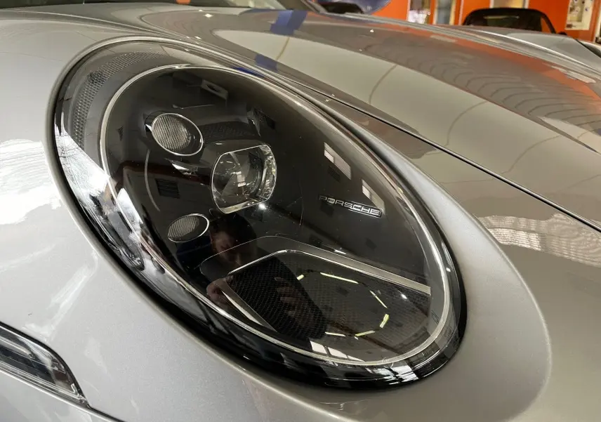 Gros plan sur le phare avant gauche argent GT d'une Porsche 911 (992) Carrera S avec détails lumineux et logo Porsche.