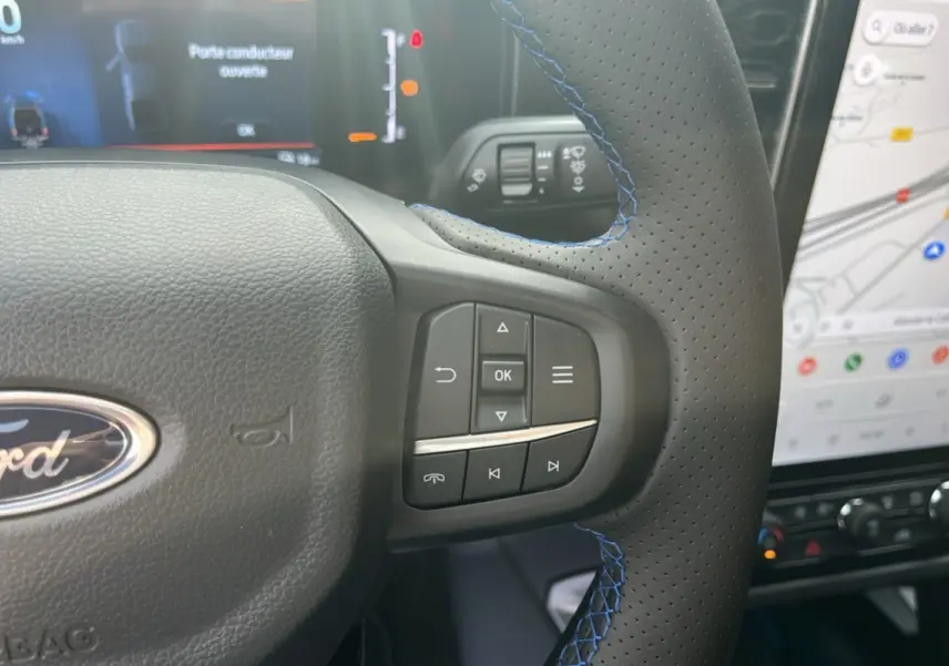 Gros plan sur le volant cuir perforé avec surpiqûres bleues du Ford Ranger 2025, écran tactile et tableau de bord en arrière-plan.