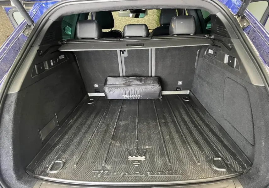 Coffre ouvert d’un Maserati Grecale bleu, vue arrière montrant tapis de coffre avec logo Trident et sac noir.