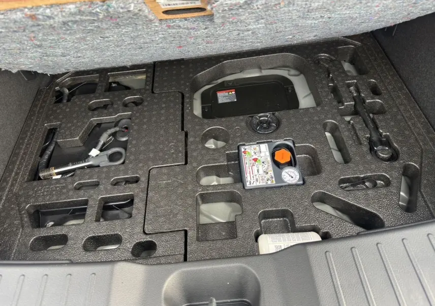 Gros plan sur le coffre ouvert d'un Nissan Juke gris squale, montrant le rangement pour outils et compresseur intégré.