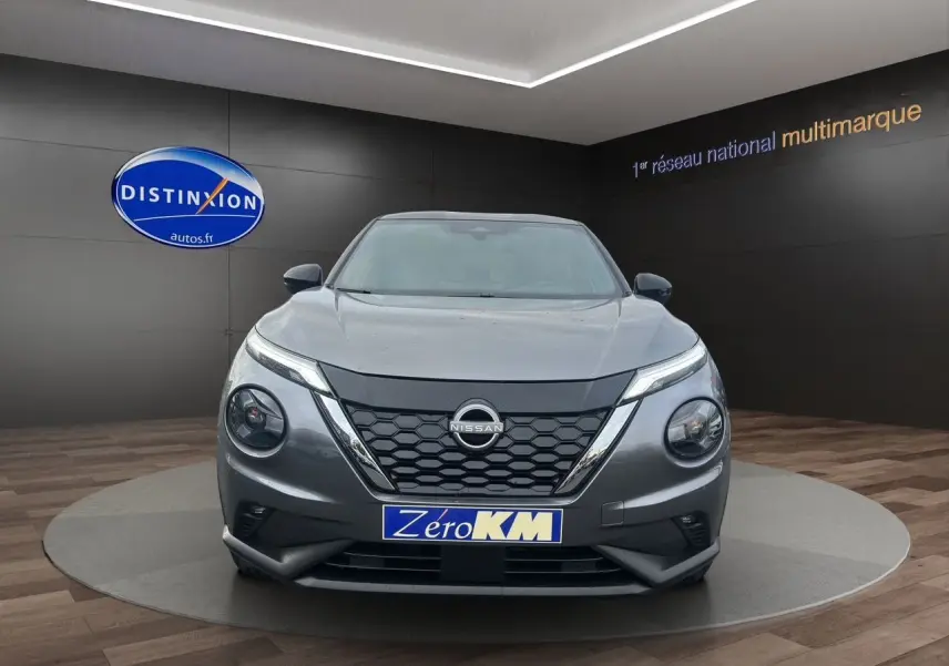 Vue frontale d'un Nissan Juke Full Hybrid gris squale, avec calandre noire et plaque "Zéro KM" en showroom.