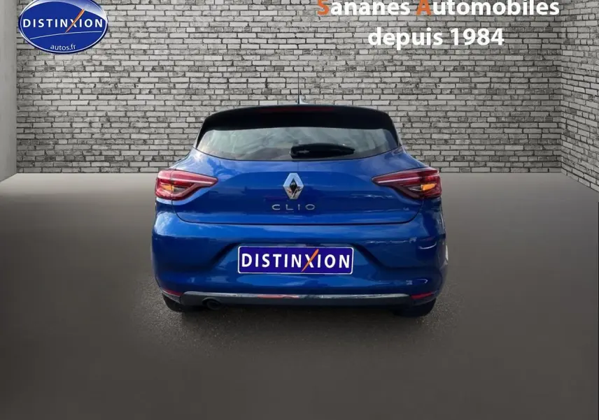 Vue arrière d'une Renault Clio 2021 bleu Iron, avec feux allumés et logo distinctif sur un fond de mur en briques.