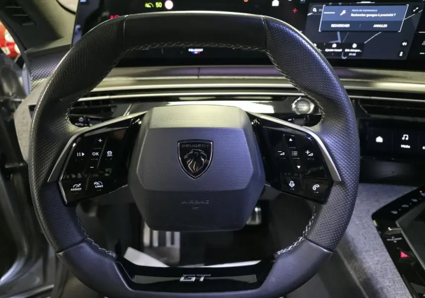 Vue rapprochée du volant cuir noir perforé avec logo Peugeot et badge GT du Peugeot 3008 gris hybride 2025.