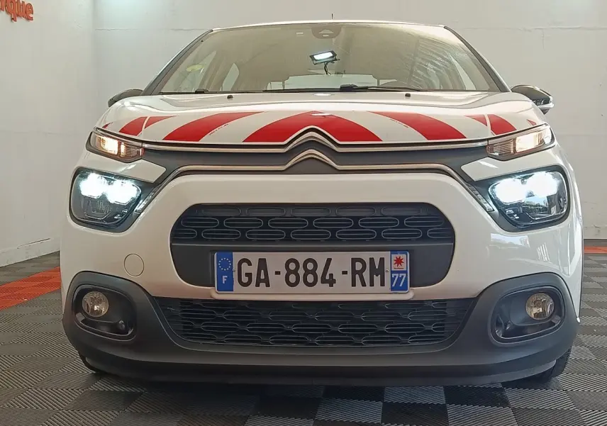 Vue frontale d'une Citroën C3 blanche avec marquage rouge sur le capot et phares allumés en intérieur.