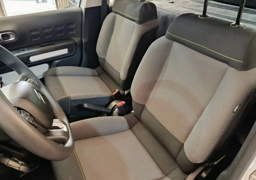 Vue intérieure côté conducteur de la Citroën C3 2021 blanche, montrant les sièges tissu gris avec surpiqûres jaunes et le volant multifonctions.