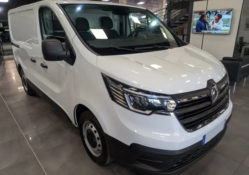 Renault Trafic Fourgon blanc minéral vu en 3/4 avant droit, avec calandre noire et phares modernes, en showroom intérieur.