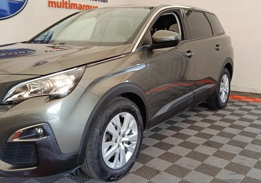 Peugeot 5008 gris foncé vue 3/4 avant droit, avec phares allumés et jantes argentées en intérieur showroom.