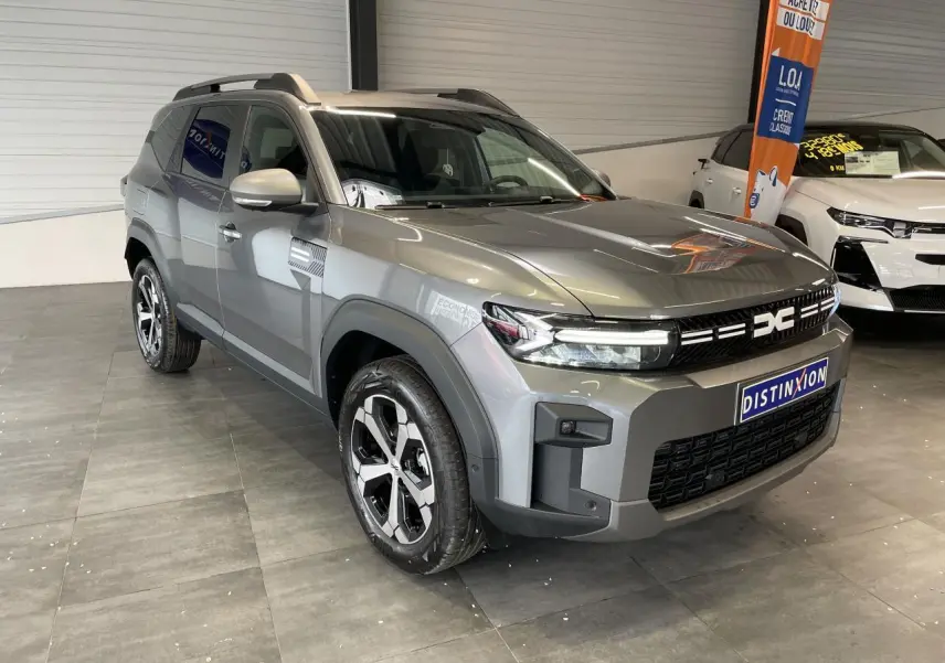 SUV Dacia Bigster gris schiste en 3/4 avant droit, avec jantes alliage diamantées et barres de toit visibles.