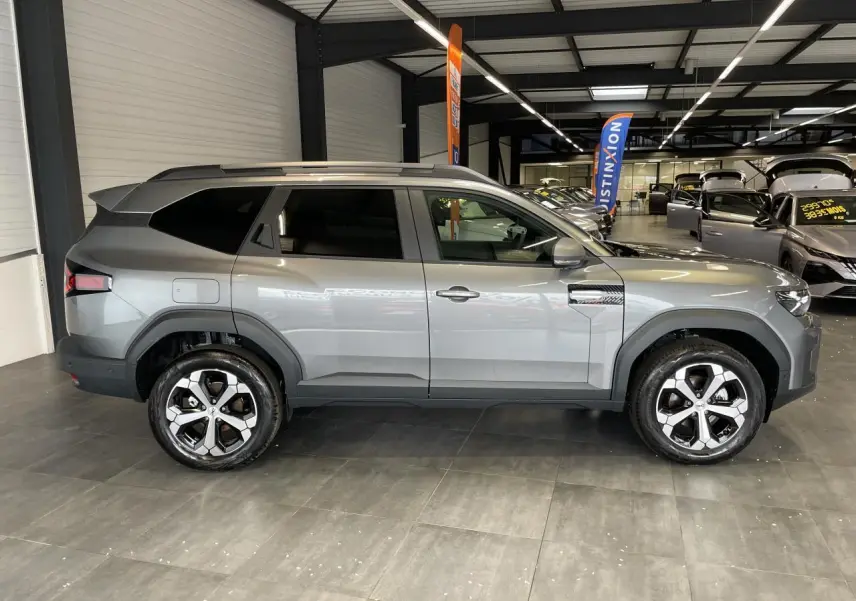 Vue de profil droit du Dacia Bigster gris schiste en showroom, avec jantes alliage 18 pouces et barres de toit longitudinales.