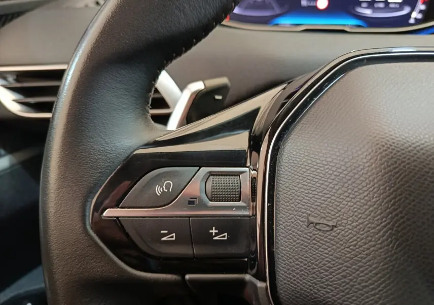 Gros plan sur les commandes vocales et volume du volant cuir noir du Peugeot 3008 2022, tableau de bord digital en arrière-plan.