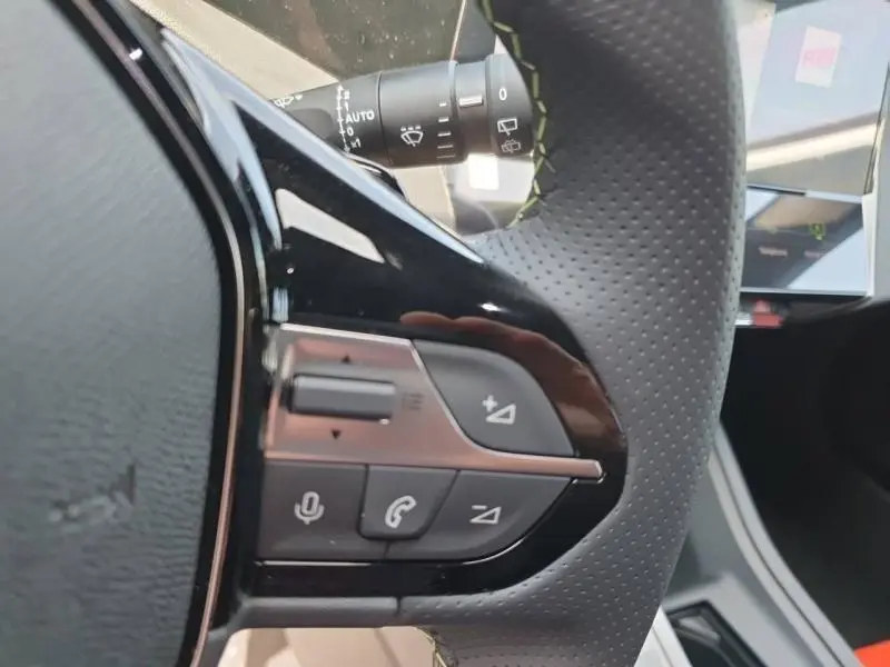 Gros plan sur les commandes gauche du volant perforé noir du Peugeot 308 Hybrid blanc Okénite, avec écran tactile en arrière-plan.