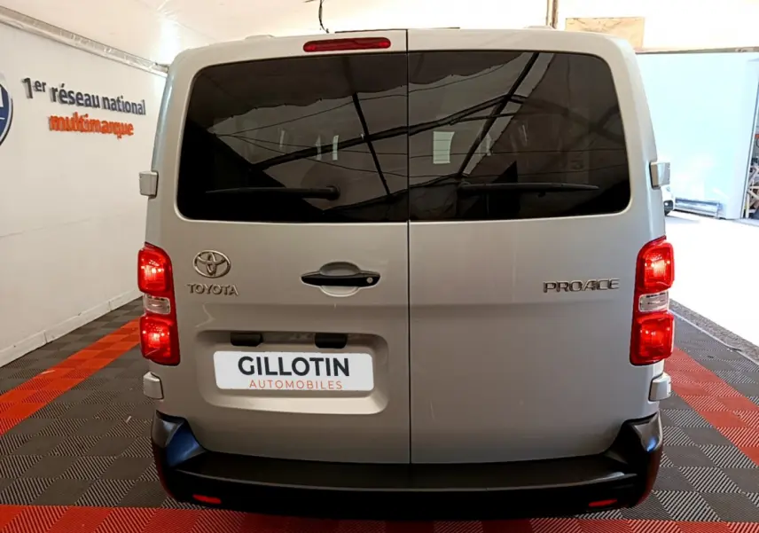Vue arrière d'un Toyota Proace Verso gris clair avec portes vitrées teintées et feux rouges allumés en intérieur showroom.