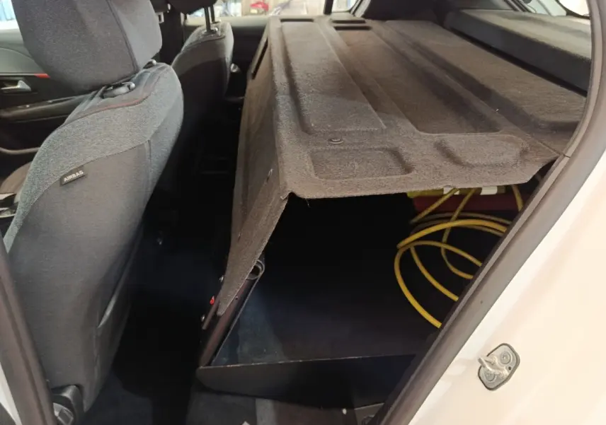 Vue intérieure côté droit arrière du coffre ouvert d'une Peugeot 208 électrique blanche, avec câble de recharge visible.