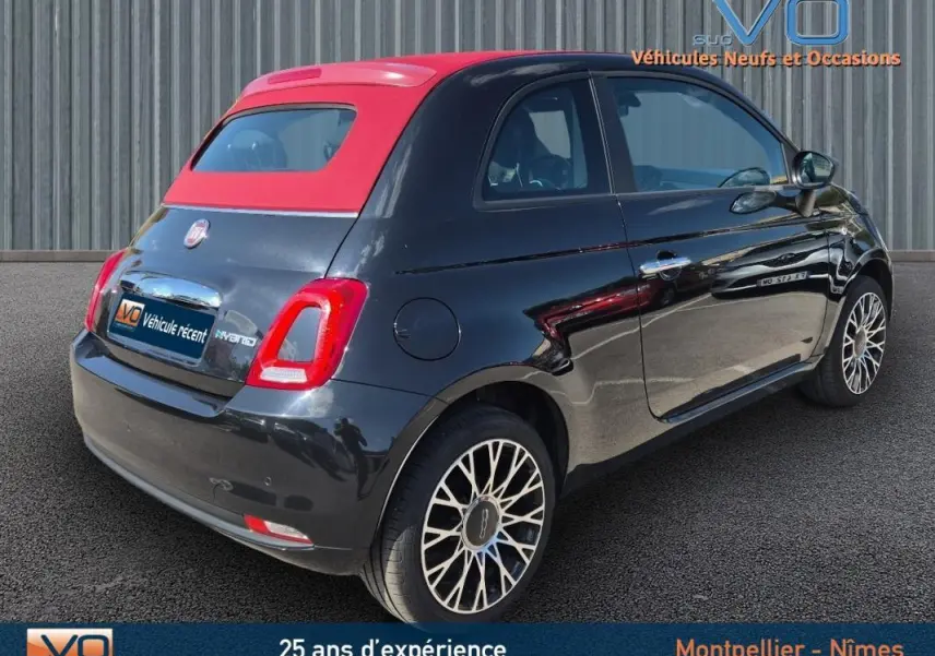 Vue 3/4 arrière droite d'une FIAT 500C noire 2024 avec toit rouge et roues alliage, version hybride essence avec roue de secours visible.