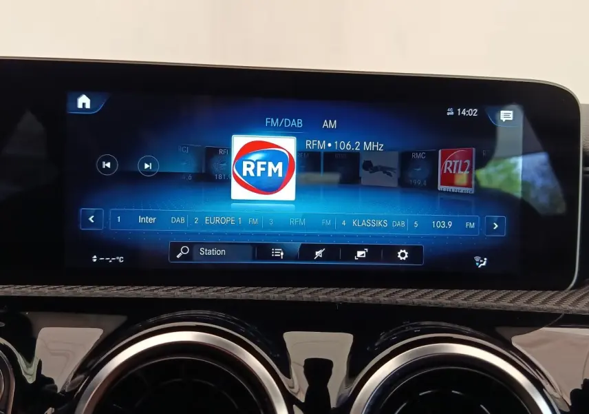 Écran tactile central affichant la radio RFM dans l'habitacle d'une Mercedes Classe A gris clair, vue de face.