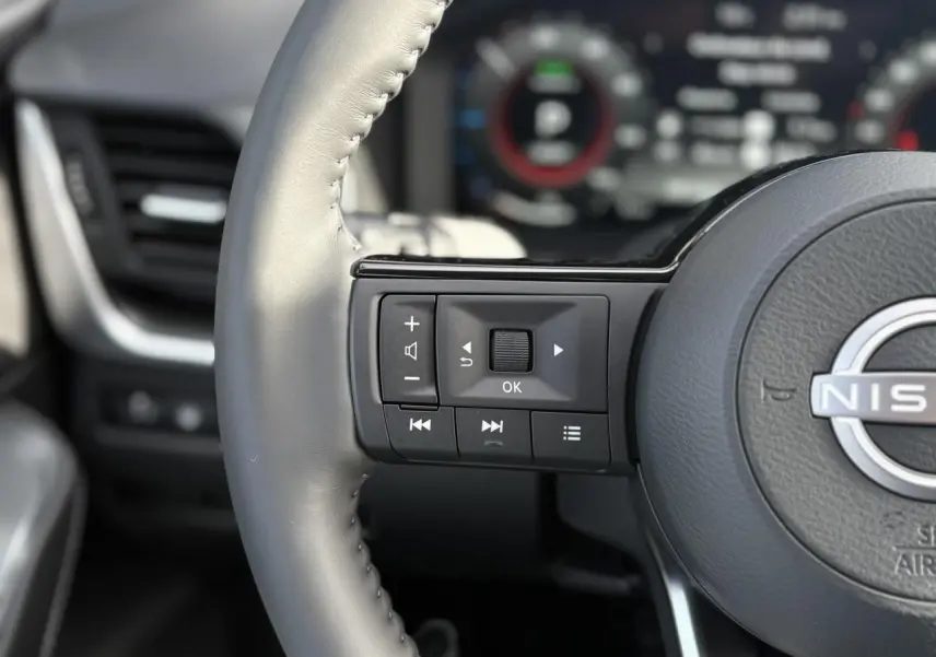 Gros plan sur les commandes gauche du volant cuir gris du Nissan Qashqai blanc nacré 2025 avec tableau de bord numérique flou.