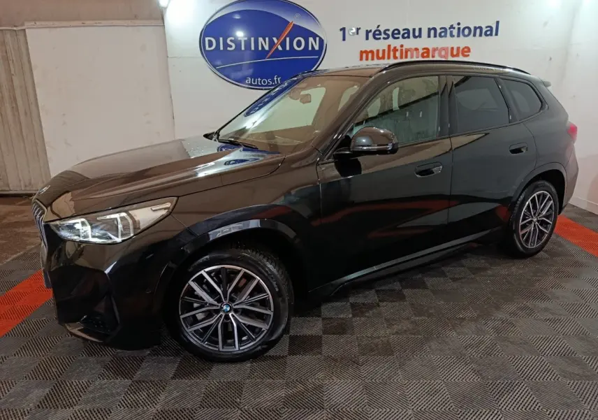 BMW X1 xDrive23i M Sport noir vue 3/4 avant droit dans un garage avec sol à damier et logo Distinxion en arrière-plan.