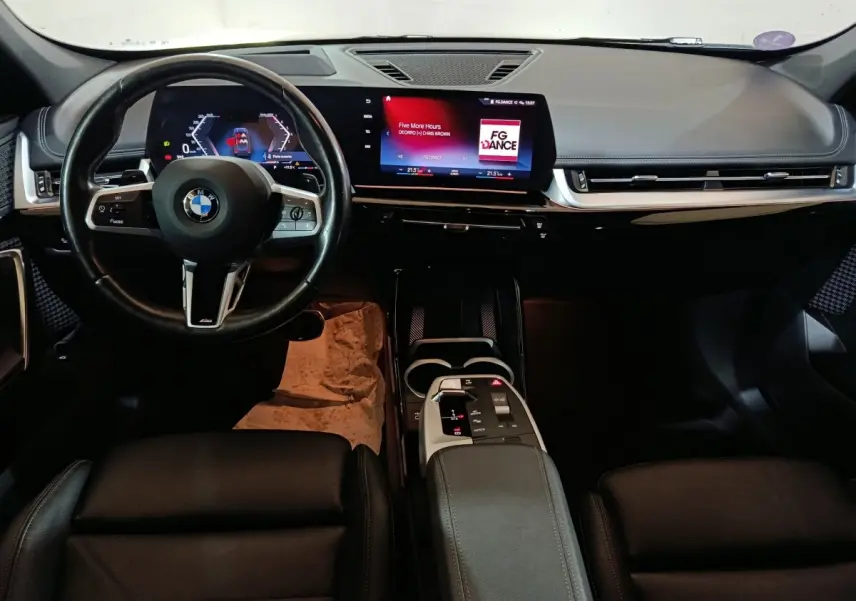 Vue intérieure avant du tableau de bord noir du BMW X1 xDrive23i M Sport avec écran tactile et volant cuir multifonctions.