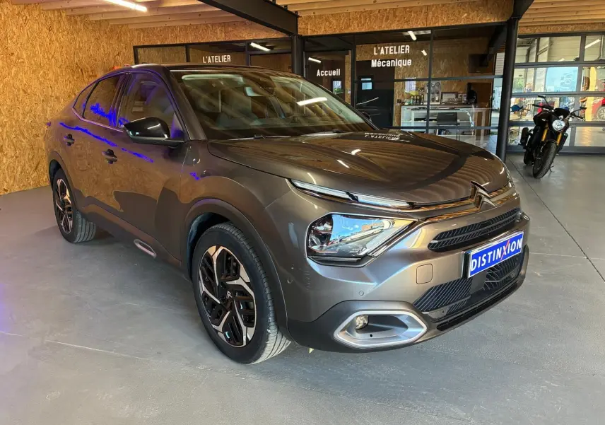 Citroën C4 X gris en 3/4 avant droit dans un atelier, avec jantes noires et éclairage LED visible.