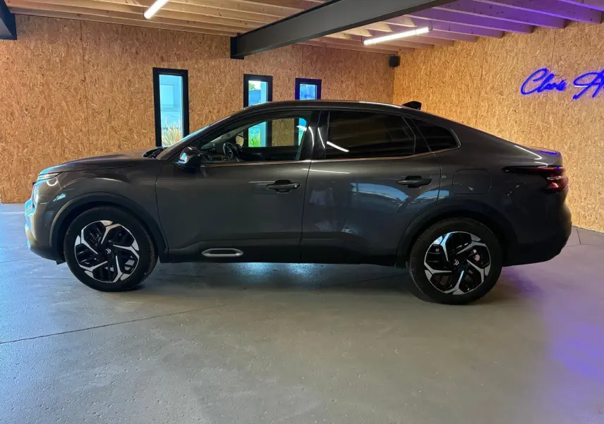 Vue de profil côté gauche d'une Citroën C4 X gris foncé 2024 avec jantes noires et vitres teintées dans un intérieur en bois.