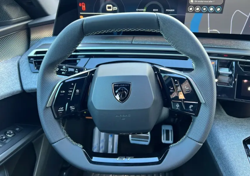 Vue rapprochée du volant cuir gris perforé avec logo Peugeot et inscription GT, tableau de bord numérique en arrière-plan.