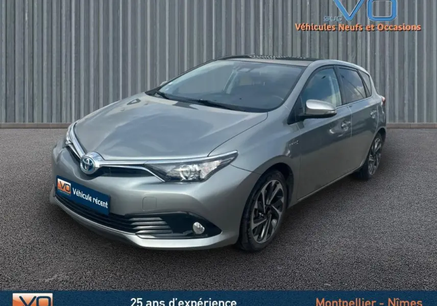 Toyota Auris Business Hybride gris clair vue 3/4 avant droit, avec jantes alliage 17 pouces et phares antibrouillard.