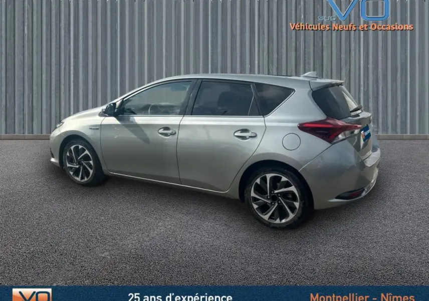 Profil côté gauche d'une Toyota Auris Business Hybride gris clair de 2016 avec jantes alliage 17 pouces sur fond urbain.