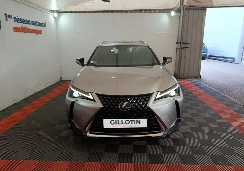 Vue avant d'un Lexus UX 250H gris clair avec calandre noire en losange et phares LED allumés dans un showroom.