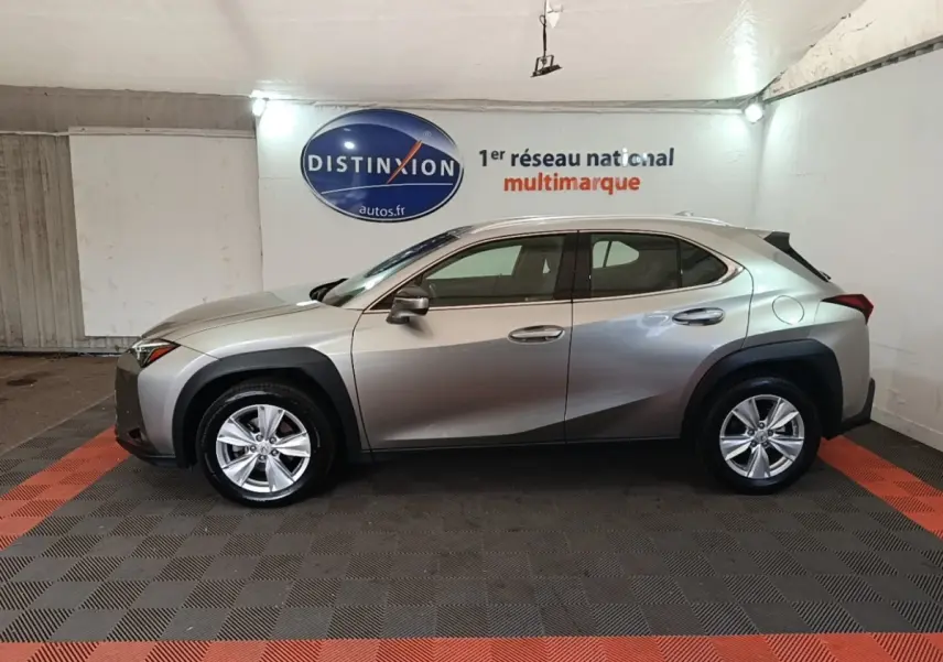 Profil droit d'un Lexus UX 250H gris clair avec protections noires autour des passages de roues, en intérieur showroom.