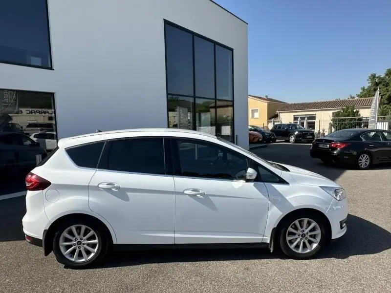 Ford C-MAX blanc vue profil côté gauche, stationné devant un bâtiment moderne sous un ciel bleu clair.