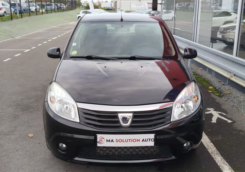 Vue avant d'une Dacia Sandero noire de 2011, avec calandre chromée et phares allumés, stationnée devant un showroom.