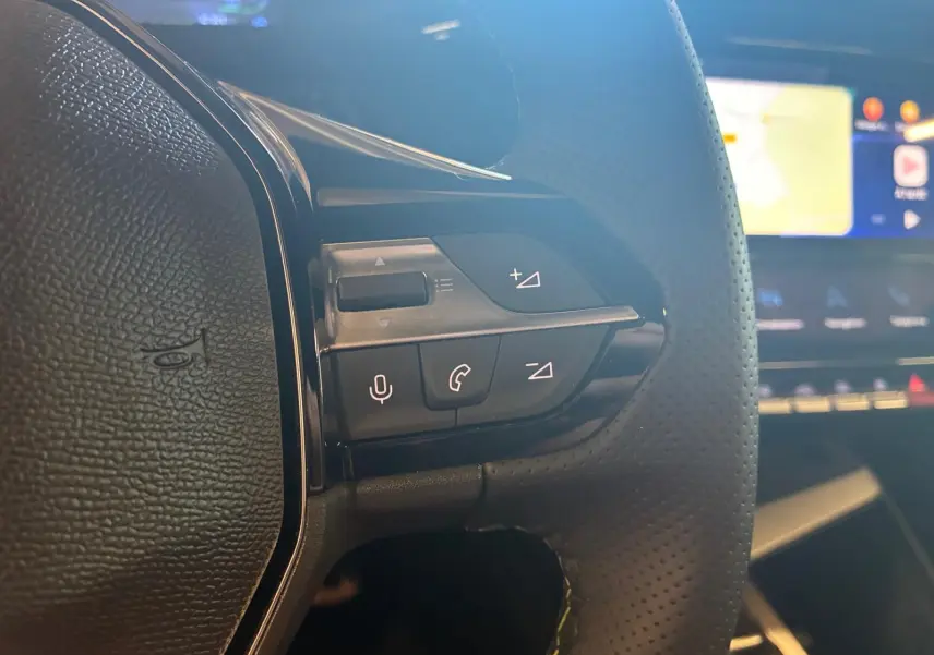 Gros plan sur les commandes gauche du volant perforé noir de la Peugeot 308 gris, avec écran tactile flou en arrière-plan.