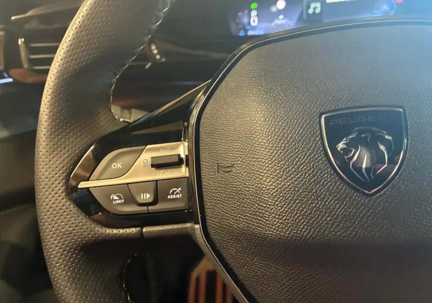 Gros plan sur le volant noir perforé de la Peugeot 308 1.2 HYBRID 136 GT avec commandes intégrées et logo lion central.