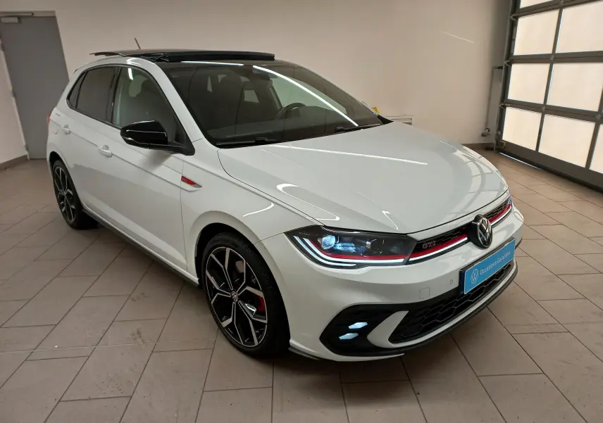 Volkswagen Polo GTI blanc pur avec toit noir, vue 3/4 avant droit, phares allumés et jantes sport noires.