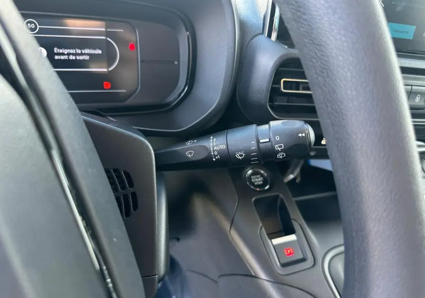Prise 12V noire sur console centrale côté passager du Citroën Berlingo III blanc, plancher moquette visible.