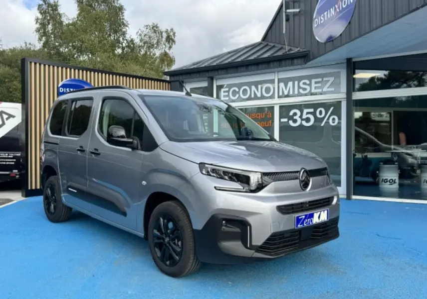Vue 3/4 avant gauche d'un Citroën Berlingo gris acier avec jantes noires, stationné devant un magasin.