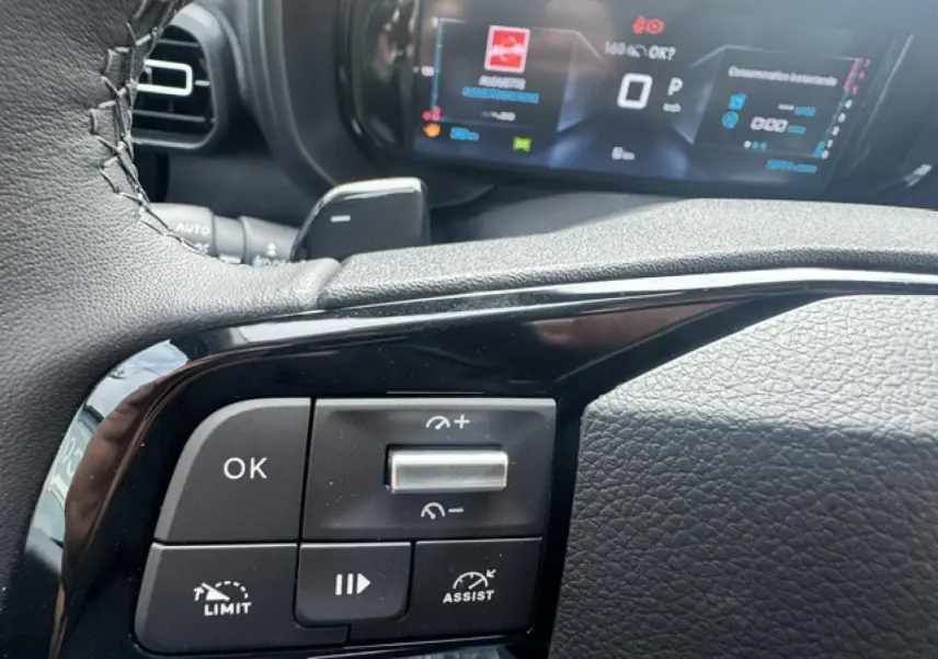 Gros plan sur les commandes au volant cuir noir du Citroën Berlingo 2026, avec affichage numérique en arrière-plan.