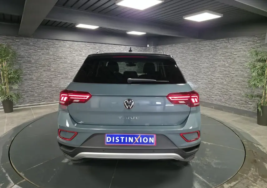 Vue arrière d’un Volkswagen T-Roc bleu métallisé 2025 avec toit noir et feux arrière LED allumés dans un showroom.