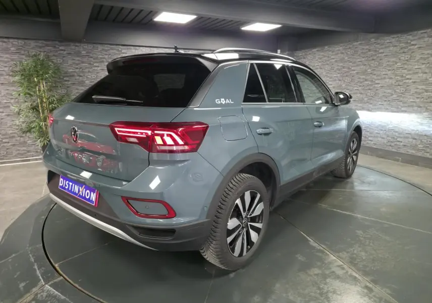 Vue 3/4 arrière droit du Volkswagen T-Roc bleu avec toit noir et feux arrière LED allumés en intérieur.