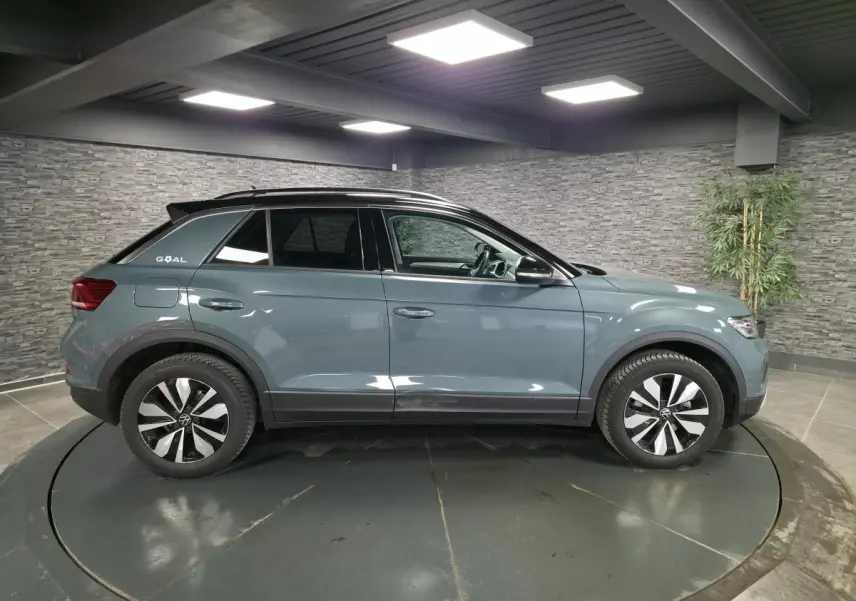 Vue de profil côté gauche d'un Volkswagen T-Roc bleu métallisé avec toit noir et jantes distinctives en intérieur showroom.