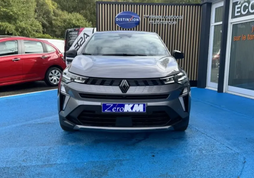 Vue avant d'une Renault Symbioz gris Cassiopée métallisé stationnée sur un sol bleu devant un bâtiment commercial.