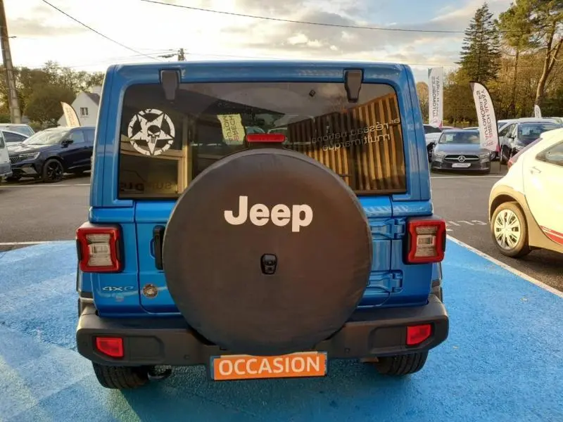 Vue arrière d'une Jeep Wrangler Hydro Blue avec roue de secours noire et plaque orange "OCCASION" sur parking.